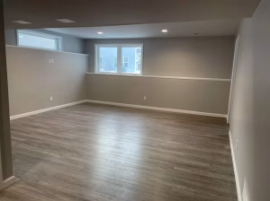 Empty basement room with gray walls, wood-like flooring, two windows, and recessed ceiling lights.