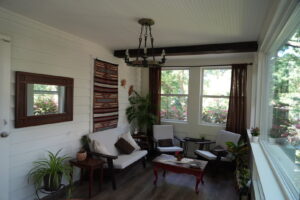 A sunlit sitting room with white walls, several chairs, houseplants, and a large window overlooking greenery.
