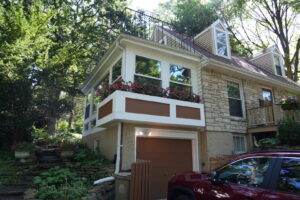 Two-story stone house with large windows, a garage, flower boxes, and surrounded by dense trees and greenery.