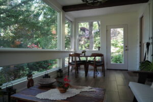 Sunlit enclosed porch with a table and chairs, wooden floor, and large windows overlooking a wooded yard.