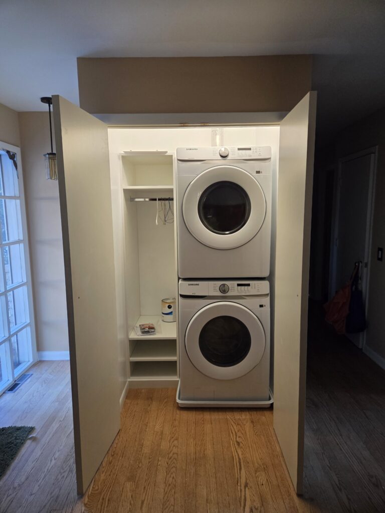 Stacked washer and dryer in a closet with open doors, shelves, and laundry supplies on a wooden floor.