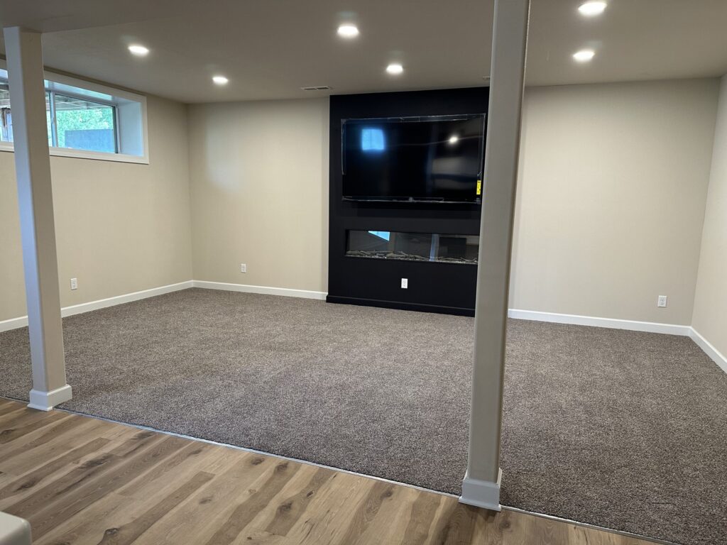 Basement room with beige walls, carpeted floor, two support columns, wall-mounted TV, and electric fireplace.